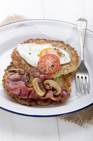 potato pancake with grilled bacon, fried egg, tomato, mushrooms and crushed pepper on an enamel plate