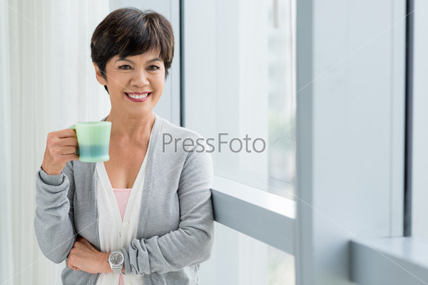 Happy smiling aged woman with a cup of tea standing at the window
