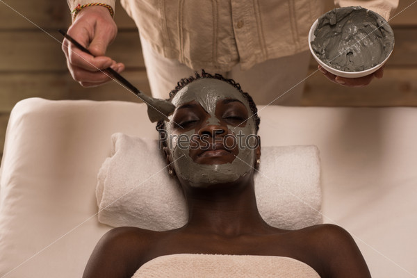Professional applying mud mask to african woman at spa salon. Top view
