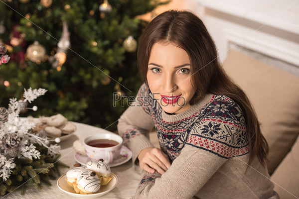 Beautiful woman wearing winter outfit drinking tea with candy at home near Christmas tree