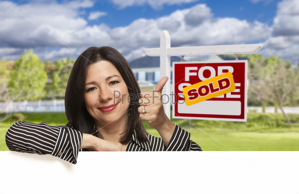 Pretty Hispanic Woman Leaning on White with Thumbs Up in Front of Beautiful House and Sold For Sale Real Estate Sign.