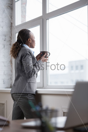 Pretty african businesswoman standing near window and drinking coffee or tea while resting and thinking about something
