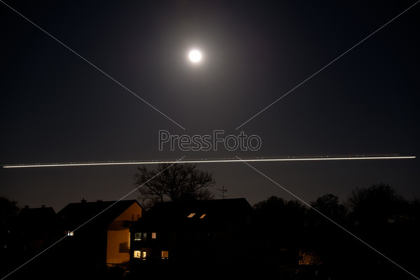 Bright moon shining over houses and trees with traces of a landing airplane