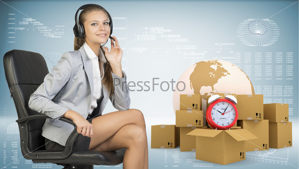 Businesswoman in headset, sitting on office chair, her hand on microphone, sitting, looking at camera, smiling. Beside are globe, commodity boxes and alarm-clock on top of one. Graphs and other