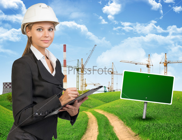 Businesswoman in hard hat standing on the road among green hills, writing on clipboard, looking at camera. Further off blank green billboard out of upright. Cranes and smoke-stacks as backdrop, stock photo