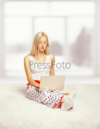 Young blonde woman in pyjamas on white whole-floor carpet browsing laptop  near window