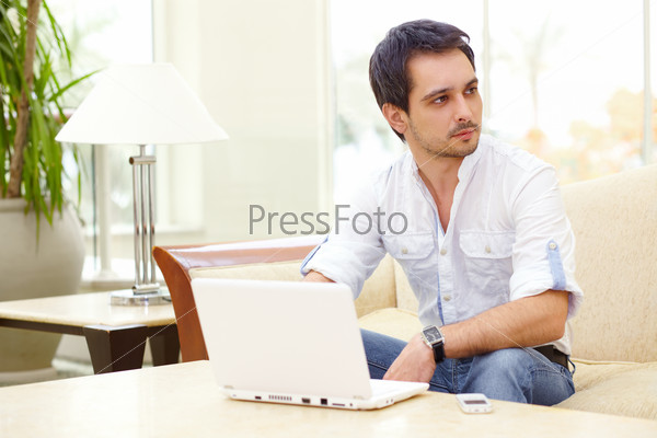 Serious young man sits waiting with white laptop in the hotel lobby or coffee shop
