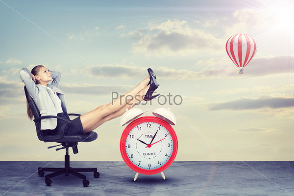 Businesswoman sitting in office chair with her hands clasped behind her head and her feet up on alarm-clock. Sky with clouds and air balloon as backdrop
