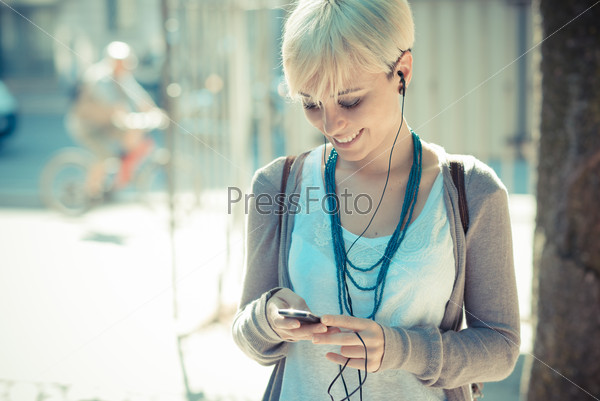 Beautiful young blonde short hair hipster woman listening music earphones in the city, stock photo