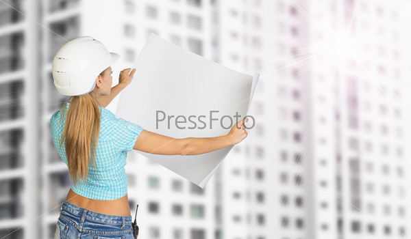 Woman in white helmet standing backwards and holding white paper sheet. Looks at paper. Building with windows as backdrop