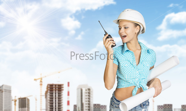 Woman talking on portable radio, holding roll of paper. Tower cranes and buildings in background