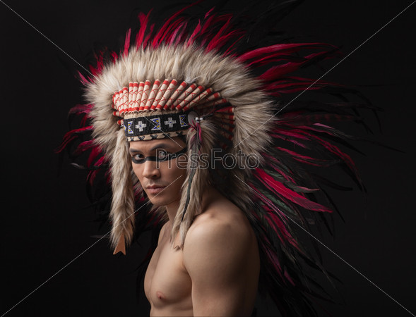 Naked indian strong man with traditional native american make up and headdress looking at the camera. Studio shot