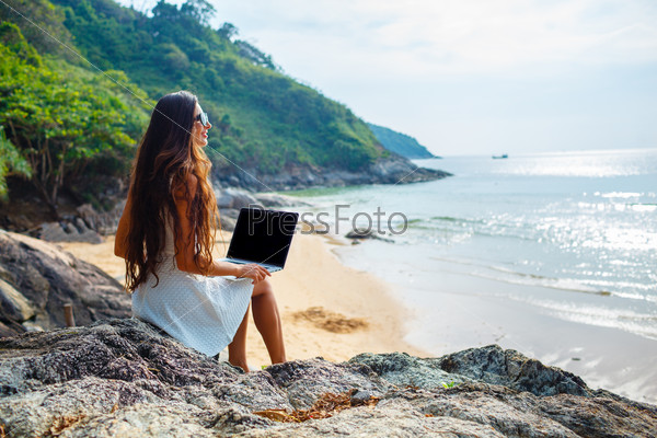 brunette woman sitting on the rock with laptop near  sea
