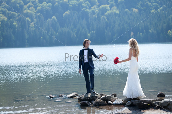 Married couple having a relaxing and fun moment at the lake. The groom dares his bride to follow his steps on some rocks.