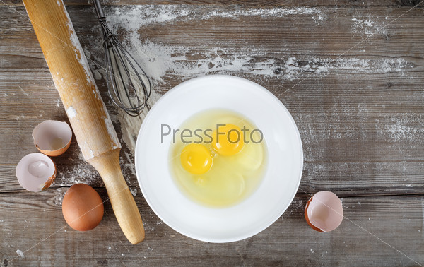 Cooking background with eggs, raw eggs in a dish, eggshells, flour, rolling pin and whisk on a table. Wooden background. Top view.
