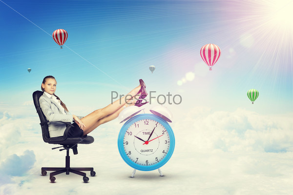 Businesswoman sits in chair, looking at camera. Put your feet up on big red alarm clock. Sky with clouds and air balloons as backdrop