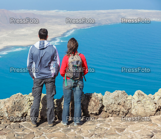 Back view sport couple with backpack standing on cliff\'s edge and looking to a island.