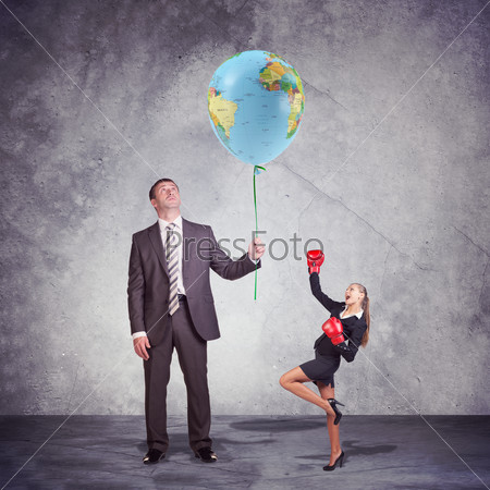 Conceptual Businessman Holding and Looking at the Balloon, with World Map Print, with a Small Businesswoman Posing in Boxing Gloves on the Side. Captured in Studio with Weathered Wall Background