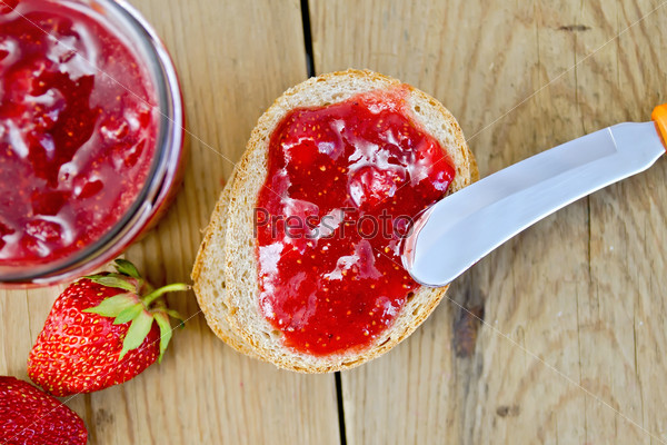 Bread with strawberry jam, a jar of jam, knife, strawberries on the background of the old wooden boards