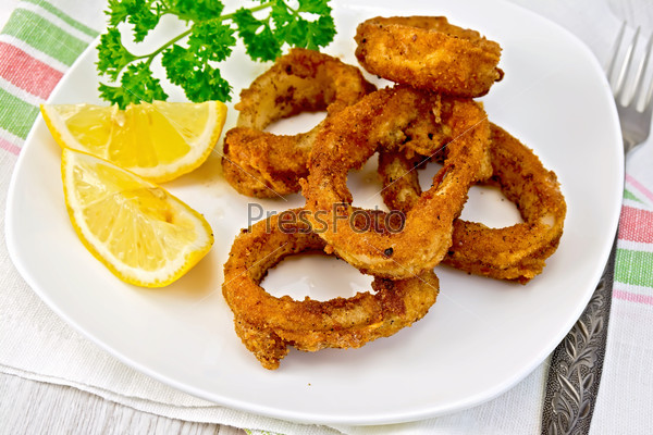 Crispy fried calamari rings on a plate with slices of lemon and parsley on a napkin on the background of wooden boards