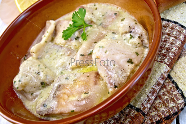 Fish stew with cream sauce in a ceramic pan, lemon, parsley, napkin on background light wooden boards