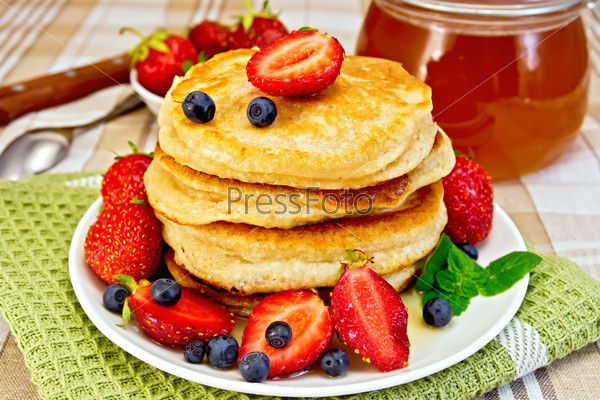 A stack of pancakes with strawberries, blueberries and honey, a jar of honey, doily on linen tablecloth background