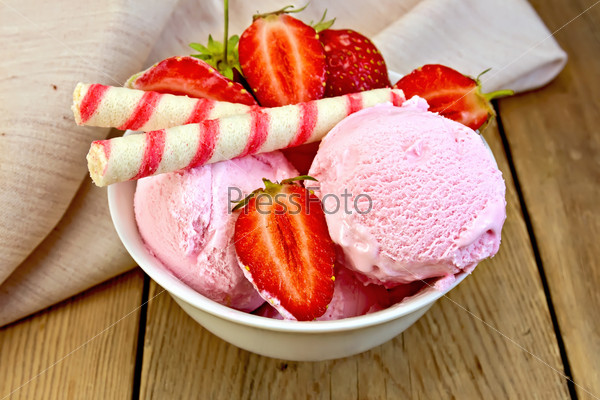 Strawberry ice cream in a white bowl with strawberries and wafer rolls, napkin on a wooden boards background