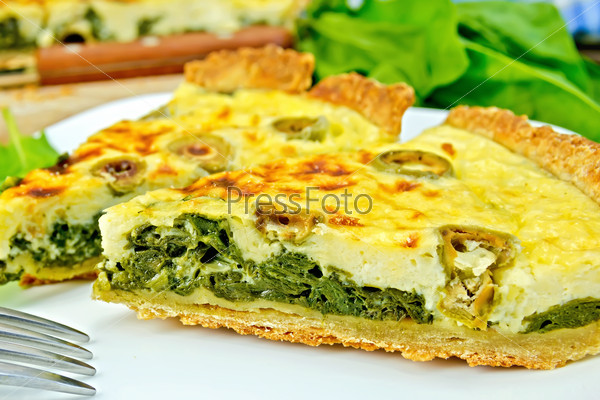 Two pieces of pie with spinach, cheese and olives on a plate, spinach leaves, knife, fork on the background of wooden boards