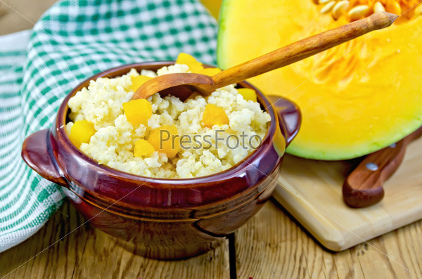 Millet porridge with pumpkin and a wooden spoon in ceramic ware, cut raw pumpkin, doily on a wooden boards background
