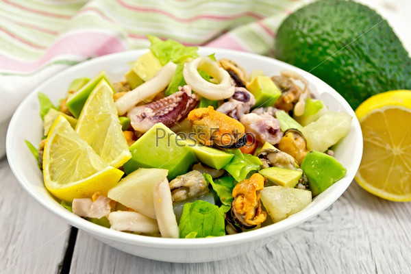 Salad with shrimps, octopus, mussels and calamari with avocado, lettuce, lemon and pineapple on a plate, napkin on the background light wooden boards