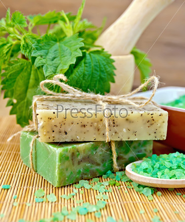 Two bars of homemade soap with twine, bath salt, nettle in a mortar on a wooden boards background