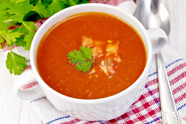 Tomato soup with pieces of vegetables in a white bowl on a napkin, spoon, tomato, parsley on a light background boards
