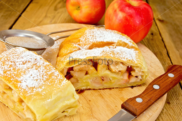 Apple strudel with icing sugar, apples, tea strainer, knife on a wooden boards background