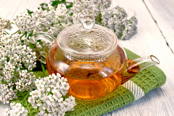 Yarrow tea in a glass teapot on a green napkin, fresh yarrow flowers on a background of pale wooden plank