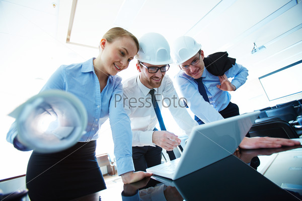 Young architect pointing at laptop display while explaining data to colleagues, stock photo