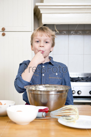Young child with sweet teeth, tasting batter from a bowl in a kitchen, wearing a blue shirt, with bowls, plates, and a whisk on the kitchen counter in front of him