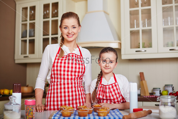 Pretty girl and her mother looking at camera with baked muffins on table near by