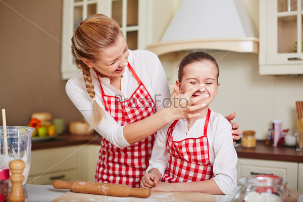 Laughing girl and her mother in aprons having fun in the kitchen