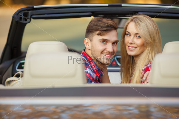 handsome couple looking at camera sitting in a car, view from rear seat in convertable car.  Lifestyle with beautiful cheerful lovers.