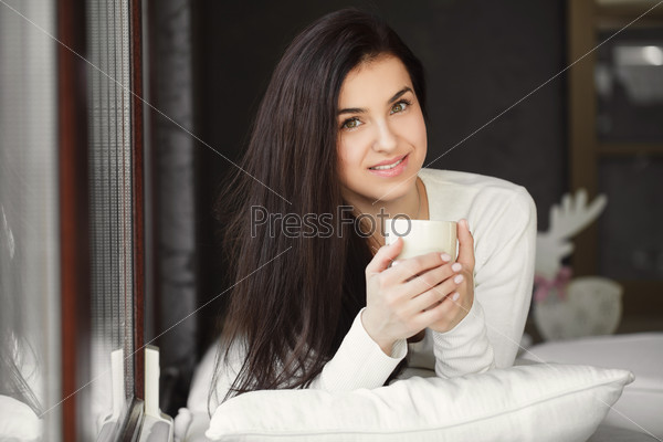 Attractive woman with a cup of coffee on the bed. Beautiful Girl Drinking Tea sitting in front of the window Indoor. Woman with the Cup of Hot Beverage.