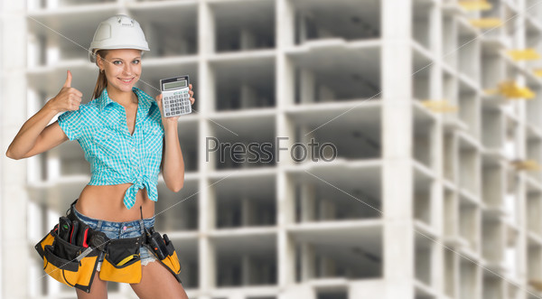 Young woman in hard hat holding calculator and looking at camera. Industrial background