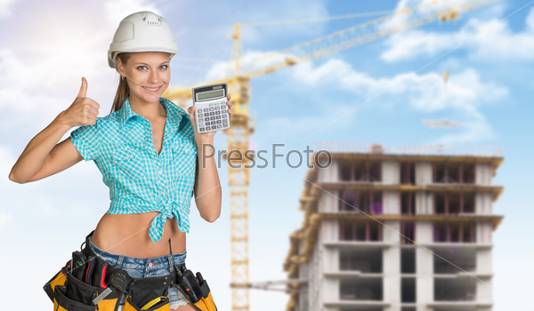 Smiling young woman in hard hat holding calculator and looking at camera. Industrial background