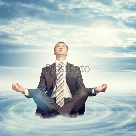 Businessman sitting in lotus position on cloud with circles and looking up on blue sky background, stock photo