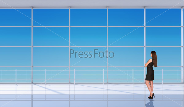 Young businesslady half-turned in black dress with crossed arms and standing back in front of window. Interior view