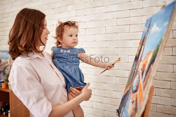 Young mother and her cute little daughter with paintbrushes looking at painting