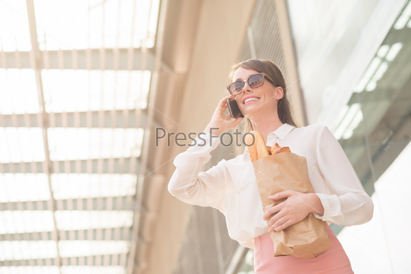 Cheerful stylish woman holding shopping bag with groceries and calling on the phone
