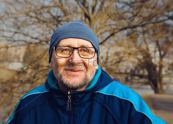 portrait of a smiling elderly man wearing glasses, a gray hat and a blue jacket in the city park