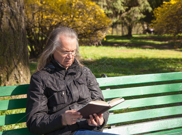 haired elderly man reading a book sitting on a bench in city park