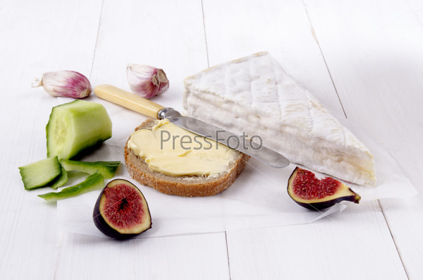 french camembert, slice bread with butter, fig, garlic, knife and peeled cucumber on white paper