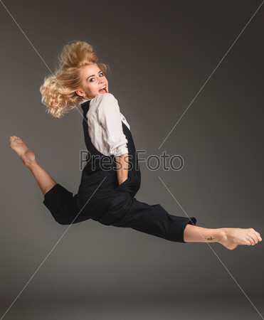 Beauty blond woman in a black suit and white shirt in ballet jump on a gray background
